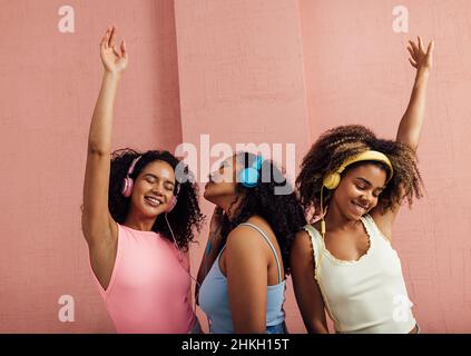 Groupe de jeunes femmes portant des écouteurs et dansant ensemble.Trois femmes en décontracté appréciant la musique contre le mur rose. Banque D'Images