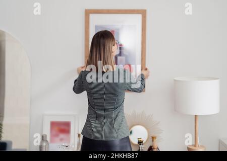 Rénovation.Vue arrière d'une femme accrochant une photo sur un mur blanc dans un intérieur lumineux Banque D'Images