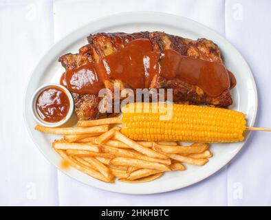 Costelinha de porco ao molho barbecue com batatas fritas e milho em fundo de madeira branca. Banque D'Images