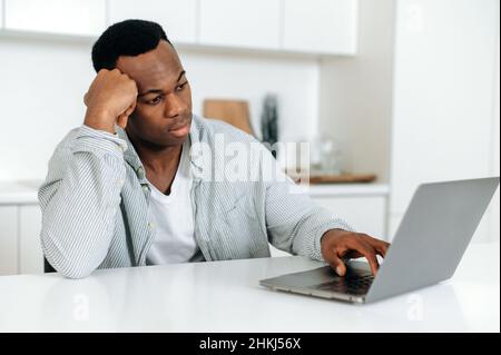 Ennuyé frustré afro-américain gars, indépendant, designer, assis à une table à la maison, vêtu de vêtements décontractés, travaillant à un ordinateur portable, fatigué, dans les besoins de repos ou de pause, contrarié Banque D'Images