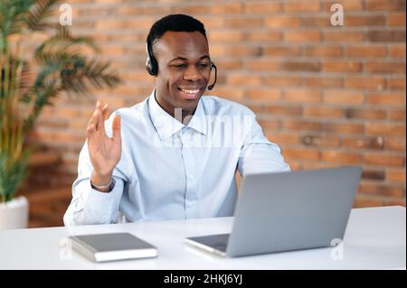 Positif attrayant Afro-américain gars dans le casque, opérateur de ligne d'assistance, employé de soutien, consultant en ligne assis à un ordinateur portable dans le bureau, travaille avec un client sur appel vidéo en utilisant le casque, salutations Banque D'Images