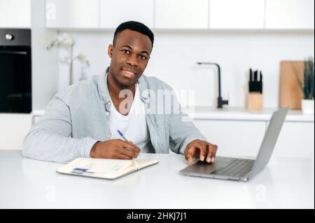 Attrayant Afro-américain gars, étudiant, indépendant, assis à une table dans la cuisine, utilise l'ordinateur portable et le bloc-notes pour l'apprentissage en ligne, regarde la caméra, écoute une conférence, prend des notes, sourire amical Banque D'Images