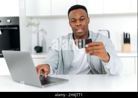 Jeune homme afro-américain positif, assis à une table dans la cuisine, en utilisant un ordinateur et une carte bancaire pour payer les achats et les livraisons en ligne, en saisissant des données, en regardant une carte, en souriant. Banque D'Images