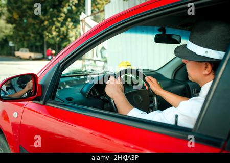 Jeune homme dans un chapeau conduisant une voiture.Sur le tableau de bord de la voiture se trouve un bouquet de fleurs blanc-jaune. Banque D'Images