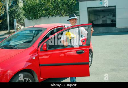 Un jeune homme dans un chapeau sort de la voiture.Dans les mains d'un homme est un bouquet de fleurs blanc-jaune. Banque D'Images