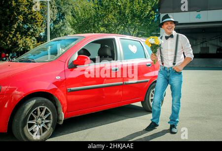 Jeune homme dans un chapeau près d'une voiture rouge.Dans les mains d'un homme est un bouquet de fleurs blanc-jaune. Banque D'Images