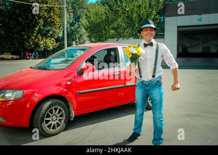 Jeune homme dans un chapeau près d'une voiture rouge.Dans les mains d'un homme est un bouquet de fleurs blanc-jaune. Banque D'Images