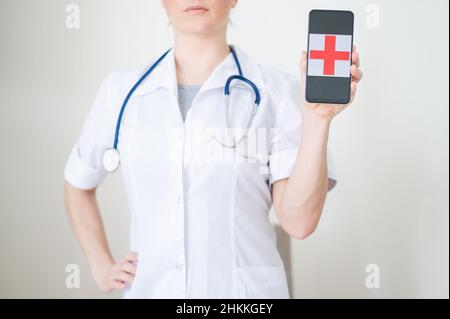 Une femme médecin sans visage tient un smartphone avec une croix rouge sur l'écran. Infirmière méconnaissable en manteau médical faisant la démonstration d'un téléphone cellulaire. Banque D'Images