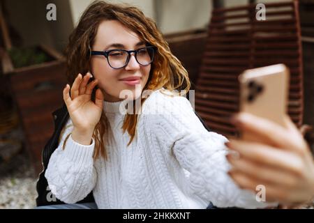 Bonne jeune caucasienne, millénaire ou la femme de génération z avec de longs cheveux de brunette streaming avec le smartphone en main, le tournage de blog de médias sociaux dans le café extérieur moderne.Hipster Influencer en utilisant les réseaux sociaux. Banque D'Images