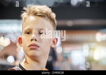 Un adolescent aux cheveux blonds ennuyé avec une expression faciale sérieuse se trouve dans la salle d'attente de l'aéroport contre une foule floue de très près Banque D'Images