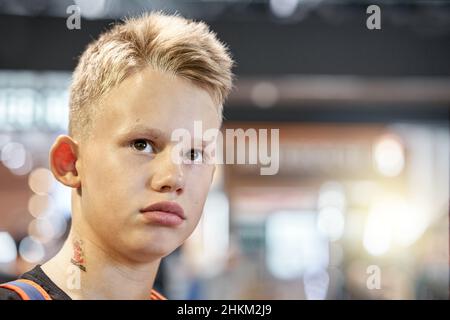 Un adolescent aux cheveux blonds ennuyé avec une expression faciale sérieuse se trouve dans la salle d'attente de l'aéroport contre une foule floue de très près Banque D'Images