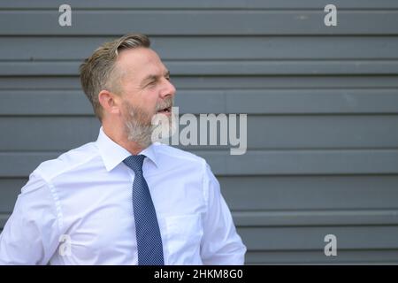 Un homme d'affaires chevronné perplexe en regardant de côté à la bouche ouverte alors qu'il se posait devant un mur gris avec des bras pliés en gros plan Banque D'Images