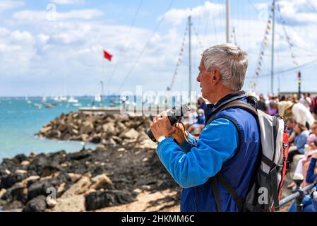 Homme regardant la course de yacht avec des jumelles pendant la semaine de Cowes avec la foule bordant le mur de mer devant le Royal Yacht Squadron, Cowes, Royaume-Uni Banque D'Images