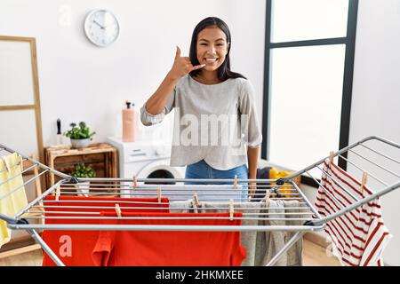 Jeune femme hispanique mettant la lessive fraîche sur la corde à linge souriant faisant le téléphone geste avec la main et les doigts comme parler au téléphone. Communicati Banque D'Images