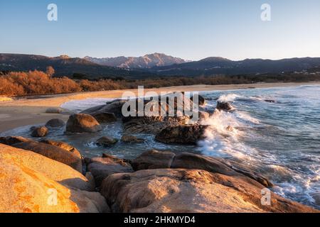 Vagues qui s'envolent sur des rochers à Areno plage près d'Algajola dans la région de Balagne en Corse avec une plage de sable déserte derrière et des montagnes enneigées i Banque D'Images