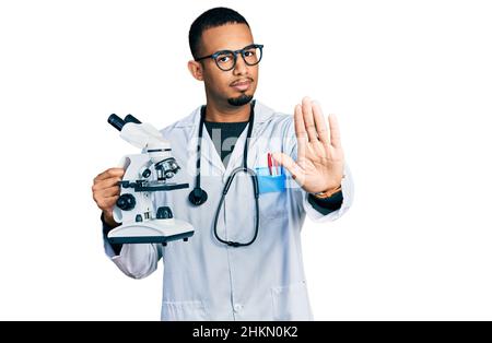 Jeune afro-américain portant un uniforme scientifique tenant un microscope à main ouverte faisant signe d'arrêt avec une expression sérieuse et confiante, la défense Banque D'Images