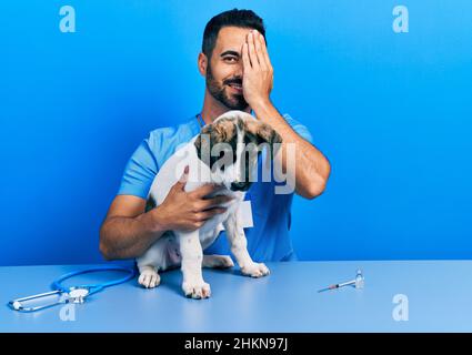 Beau vétérinaire hispanique avec la barbe de vérifier la santé de chien couvrant un oeil avec la main, sourire confiant sur le visage et émotion surprise. Banque D'Images