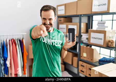 Homme d'âge moyen avec une barbe portant un t-shirt de bénévole tenant un smartphone pointant vers vous et l'appareil photo avec les doigts, souriant positif et gai Banque D'Images