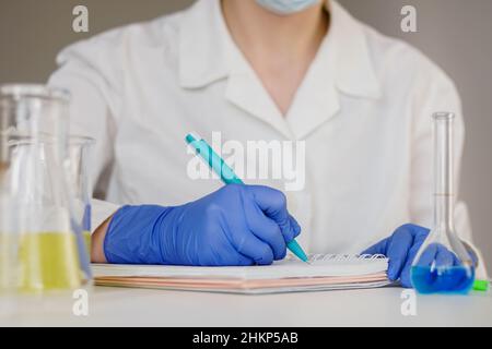 Une femme pharmacienne en gants bleus écrit les résultats de la recherche dans un carnet. Banque D'Images