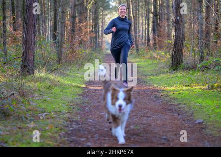 Smiling woman jogging en plein air avec son chien en avance sur un sentier de terre dans la forêt ou le parc. Banque D'Images