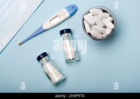 Thermomètre pour mesurer la température corporelle, masque médical, petit seau de pilules, vaccin dans un pot sur fond médical bleu. Banque D'Images