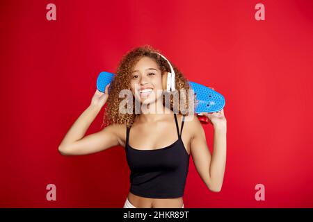 Une jolie chipster afro-américaine à poil dur avec un panneau de penny écoutant de la musique dans un casque et regardant l'appareil photo sur fond rouge en studio Banque D'Images