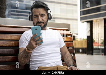 Homme hispanique avec une barbe assis sur un banc regardant son téléphone cellulaire dans un cadre urbain. Banque D'Images