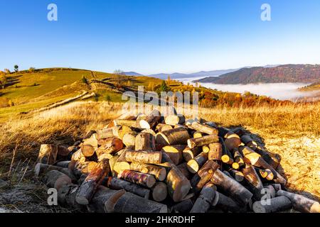 Tas de bois de chauffage haché se trouve sur l'herbe sèche dans les hautes terres contre les montagnes lointaines entourées de brouillard sous le ciel bleu au soleil d'automne coucher de soleil Banque D'Images