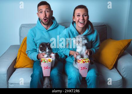Jeune couple heureux avec pop-corn et chats drôles assis sur un canapé regardant le film comique et appréciant le temps ensemble à la maison Banque D'Images
