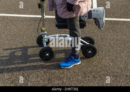 Genou scooter ou marcheur utilisé par une femme avec pied de jambe dans une chaussure chirurgicale le jour d'hiver ensoleillé. Banque D'Images