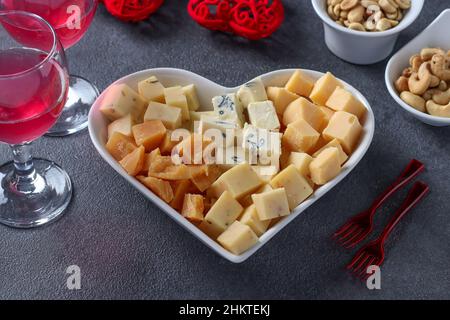 Variétés de fromage d'élite dans une assiette en forme de coeur, noix de cajou, arachides et deux verres de vin pour la Saint-Valentin sur fond gris Banque D'Images