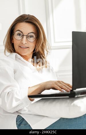 Portrait d'une femme d'âge moyen positive dans des lunettes élégantes.Belle femme en chemise de coton blanc assise sur un canapé avec ordinateur portable.Femme d'affaires sto Banque D'Images