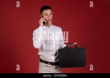 Un homme en colère dans une chemise blanche et un pantalon léger avec un sac à documents dans sa main hurle sur un téléphone portable sur le haut-parleur.Un homme stressé et déprimé sur fond rouge.Homme émotionnel Banque D'Images