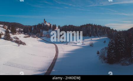 Magnifique vue aérienne de drone d'une église à Hrib Loski Potok lors d'une froide journée d'hiver ensoleillée remplie de neige.Magnifiques forêts enneigées entourant le pictu Banque D'Images