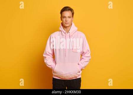 Jeune homme attentionné en sweat-shirt rose avec capuche posé sur fond jaune studio avec les mains dans la poche Banque D'Images