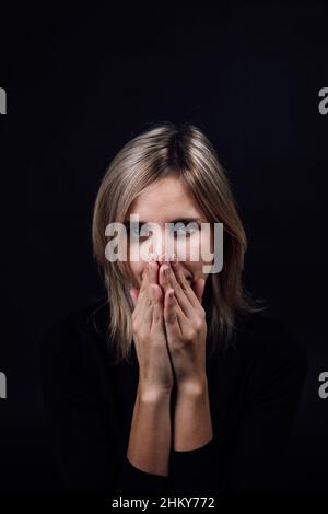 Femme effrayée avec les yeux regardant l'appareil photo, mains cachant le nez et la bouche dans un chemisier noir sur fond noir.Victime d'abus physique et psychologique Banque D'Images