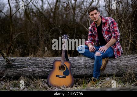 Un beau jeune homme est assis sur un arbre tombé à côté d'une guitare acoustique dans un parc naturel. Banque D'Images