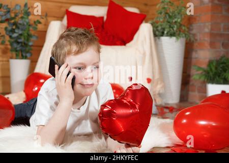 Les enfants ou les enfants de la Saint-Valentin.Petit garçon mignon avec un gadget, un téléphone portable et un ballon rouge en forme de coeur dans sa main, le jour de la Saint-Valentin.Congra Banque D'Images