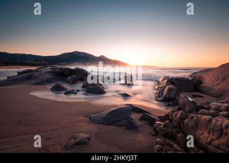 Coucher de soleil sur une crique rocheuse à Aregno Plage dans la région de Balagne en Corse avec le village d'Algajola au loin Banque D'Images