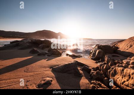 Coucher de soleil sur une crique rocheuse à Aregno Plage dans la région de Balagne en Corse avec le village d'Algajola au loin Banque D'Images