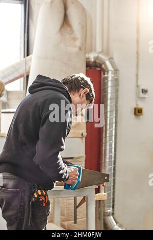 Portrait d'un charpentier formé travaillant dans son atelier de menuiserie.Menuiserie coupant une planche en bois avec une scie à main pour des meubles sur mesure.Un artisan qualifié sciant des planches de bois sur une table à l'aide d'une scie à bras portative.Tir vertical Banque D'Images