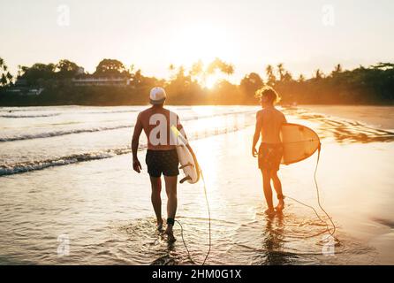 Père avec un fils adolescent marchant avec des planches de surf près de la plage de sable de l'océan avec des palmiers sur fond éclairé par le soleil de coucher de soleil.Ils sourient et ha Banque D'Images