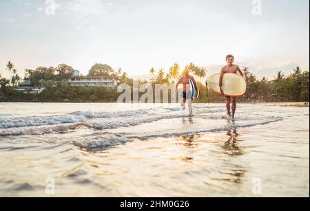 Jeune garçon adolescent avec son père portant des planches de surf alors qu'ils marchent sur la plage de sable de l'océan après avoir surfé sur fond de beau coucher de soleil.Ils sont s Banque D'Images