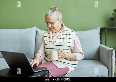Femme contemporaine sénior dans des vêtements décontractés, assise sur un canapé, prenant un verre et se connectant en réseau devant un ordinateur portable dans le salon Banque D'Images