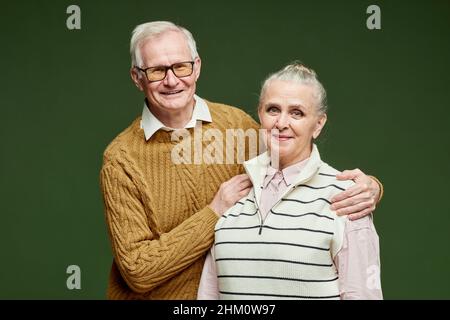 Couple senior contemporain et joyeux, en pull-overs, debout sur fond vert foncé et regardant l'appareil photo avec des sourires Banque D'Images