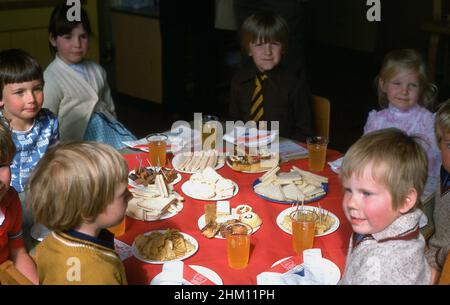 1977, historique, à l'école primaire d'un village, sandwichs et gâteaux disposés sur une table sur une nappe rouge, pendant que les enfants s'assoient pour un repas pour célébrer le Jubilé d'argent de sa Majesté, la reine Elizabeth II, Angleterre, Royaume-Uni. Banque D'Images