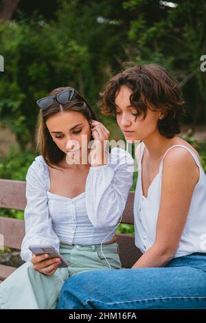 Deux jeunes filles sérieuses regardent une vidéo tout en étant assis sur un banc de bois au parc. Banque D'Images