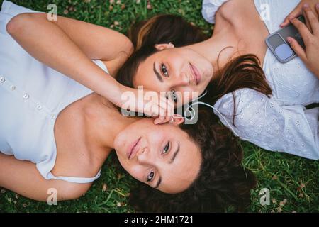 Deux adolescentes regardent l'appareil photo pendant qu'elles écoutent de la musique.L'un d'eux tient l'écouteur.Ils se détendent sur l'herbe. Banque D'Images