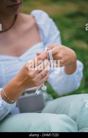 Une jeune fille enroule le câble du casque.Elle est assise sur l'herbe dans un parc. Banque D'Images