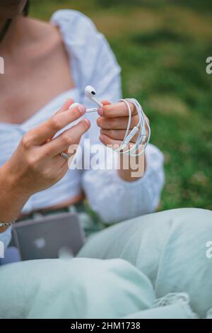 Un adolescent enroule le câble des écouteurs après avoir écouté de la musique.Elle est assise sur l'herbe dans un parc. Banque D'Images